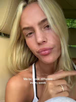 The image is a close-up selfie taken from a low angle inside a car. A fair-skinned woman with blonde hair, styled with loose waves, is the focus. She is looking towards the camera with a slightly pursed expression. She wears a white tank top, and a gold ring and delicate necklace. The primary colors are light and natural. The lighting appears to be soft, likely from an overcast day. A text overlay reads, 'Me bc look different every week.'