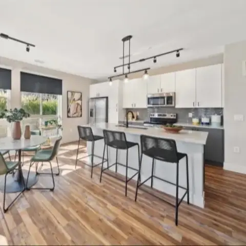 An interior shot of a modern kitchen and dining area. The kitchen features white cabinets, a gray tiled backsplash, stainless steel appliances, and a white island with black bar stools. The dining area has a round table with green chairs. The floor is hardwood, and large windows offer natural light.