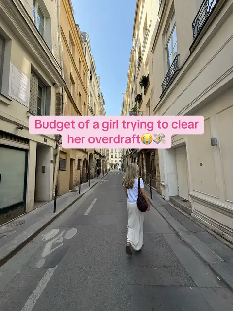 A woman in white clothing walks down a narrow European street. The buildings on either side are tall with muted yellow and beige facades. The sky is clear and blue. A text overlay in a pink rectangle sits across the top of the image.