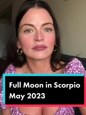 A woman with dark hair and light eyes is centered in a vertical video frame. She has light skin, rosy cheeks, and is wearing gold hoop earrings and a floral top. A black banner near the bottom of the frame has white text that reads "Full Moon in Scorpio May 2023."