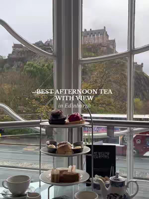 A photo taken inside a tea room with a large window overlooking a cityscape. A tiered stand with pastries and finger sandwiches sits on a table in the foreground. Outside the window, a green hill leads up to a large stone castle. The scene is bright and inviting.