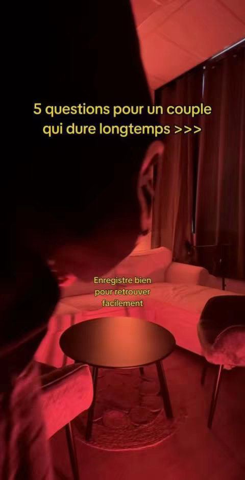 The image shows a close-up shot of a dimly lit room, predominantly red in hue. The focus is on the upper body and head of a person, partially obscuring the view, in front of a setting resembling an intimate space with a couch and a small table, the person's silhouette is visible. Text overlayed on the top is stylized in yellow font. Below, a second text is overlaid in a more subtle, smaller font.