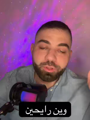 A man with short hair and a beard is centered in the frame, speaking into a microphone. He wears a light blue denim shirt, and the background is a gradient of purple and pink hues, lit with small, white, twinkling lights to give the appearance of stars. A black rectangular text box at the bottom of the frame has white text.