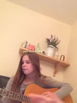 A young woman with long brown hair is sitting and playing a guitar. She is wearing a gray long-sleeve shirt. Behind her is a cream-colored wall, a wooden shelf with various items, including a small figurine, a plant in a white vase, and a candle. The text overlay says 'you didn't need "fix"'.