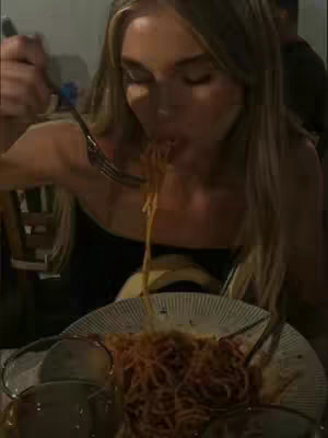 A woman eats spaghetti at a restaurant. She has long blonde hair and is wearing a black top. A plate of spaghetti sits in front of her. The photo is lit with warm lighting.