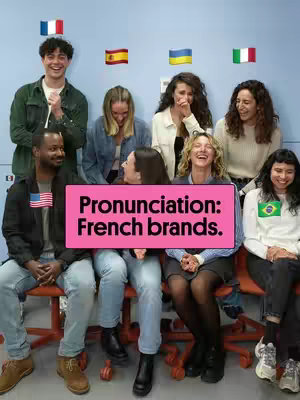 A group of diverse people are seated in front of a light blue wall. The central focus is a large, pink rectangular sign held by two people in the center of the group. Above them, several small flags are displayed. The people appear to be interacting or reacting to something outside of the frame.