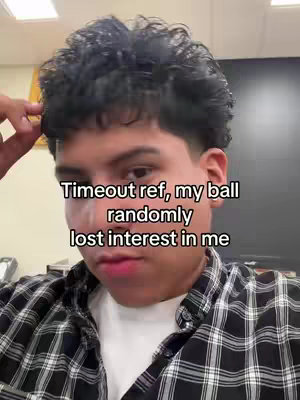 A selfie-style shot featuring a young man in a plaid shirt. The background is muted, likely inside a room, with a dark-colored surface and possibly a bulletin board visible. Text overlayed on the image reads, “Timeout ref, my ball randomly lost interest in me.”
