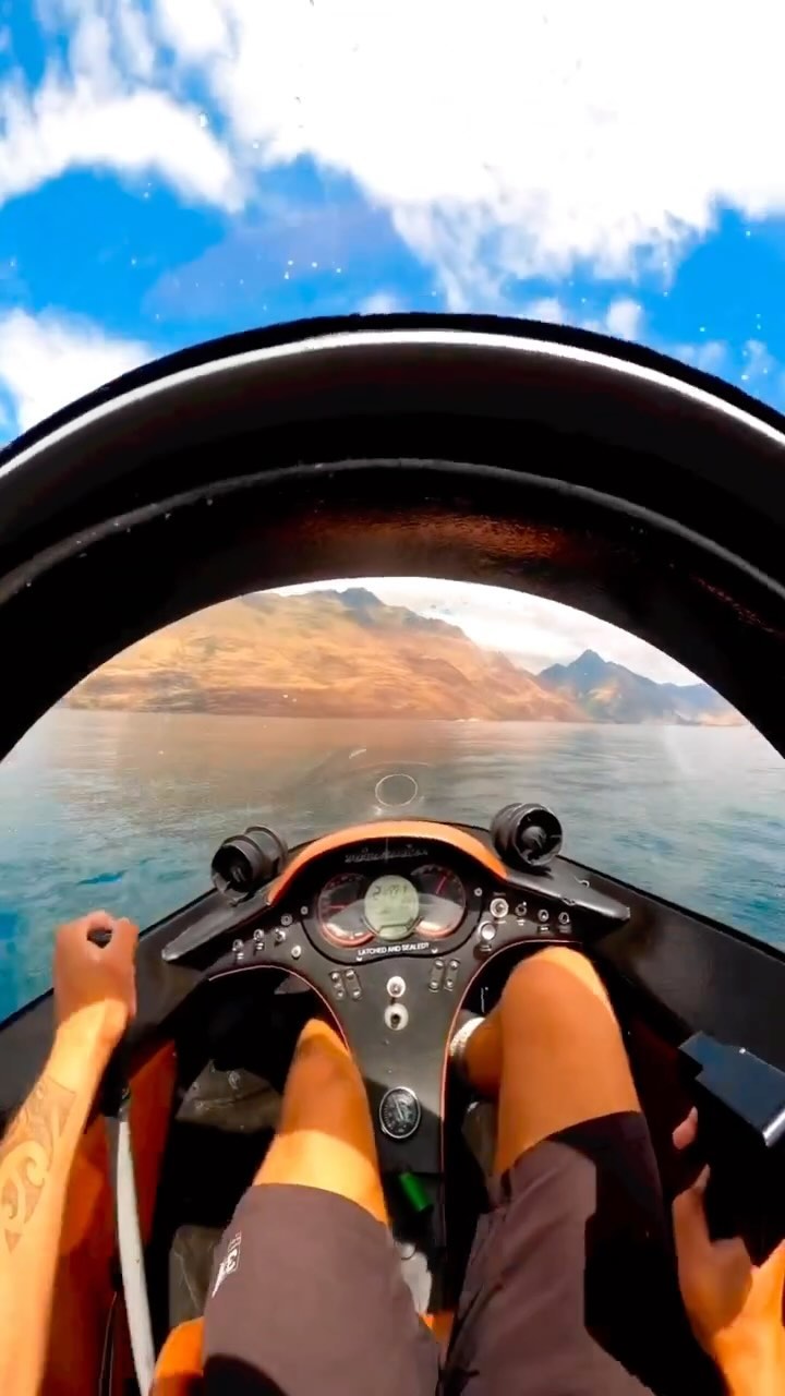 The image shows a first-person perspective from inside a small watercraft. The viewer looks out over a body of water, with a mountainous landscape under a blue sky filled with clouds. The interior of the craft is black and orange, with a control panel visible. The legs and hands of a person, presumably the driver, are also in view.