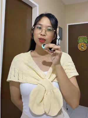 A young woman poses indoors, holding a small object up to the camera. She wears gold-rimmed glasses, a white top, and a light yellow shawl or cardigan with a knotted design. The background includes a closed brown door, hints of another door, and warm-toned walls. The composition is well-lit with a focus on the subject's face and upper body.