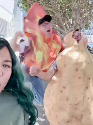 Three individuals in cosplay are positioned in front of an outdoor setting. A person on the left has long teal hair and is partially visible. In the center, a person is in a fiery costume, and on the right, a person is in a potato costume. The background includes a building on the left and greenery suggesting trees.