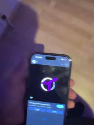 A person is holding a smartphone displaying a stylized logo with a white circle and a purple arrow. The phone screen is dark with white text and blue buttons at the bottom. The background shows a wooden floor and purple-lit surroundings.
