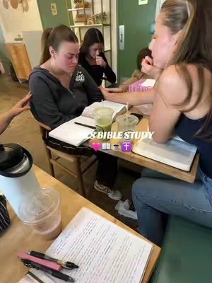 A group of young women are sitting around a table in a cafe, with open notebooks and Bibles, engaged in a Bible study session. The color palette is warm, with soft natural lighting and the scene appears to be candid.