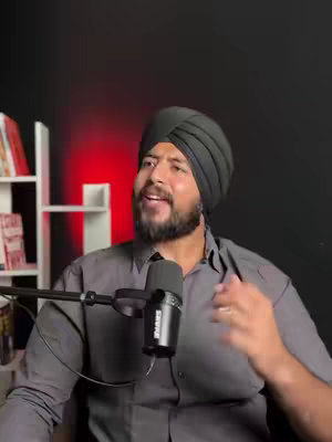 A man wearing a turban and a gray shirt is speaking into a microphone. Behind him is a dark wall with two shelves stacked with books. The lighting suggests a studio setting, with a red glow visible on the left.