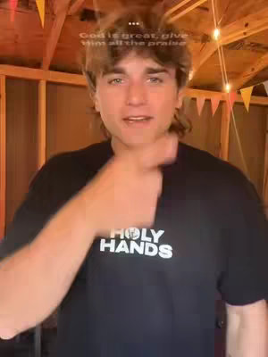 A young man with light brown hair and a mullet is centered, pointing off to the side with his right index finger. He wears a black t-shirt with white lettering in the center that reads "HOLY HANDS". Behind him is a wood structure with warm lighting from a few light bulbs. Above him is overlaid text that reads "God is great, give Him all the praise".