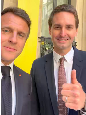 Two men in suits take a selfie in front of a yellow wall. One man, on the left, is partially visible, with his face and the top of his suit. The other man, on the right, is centered, wearing a dark suit, a white shirt, and a patterned red tie. He is giving a thumbs-up, smiling at the camera. A gold-colored ornate structure is partially visible in the background suggesting they are outside a building.
