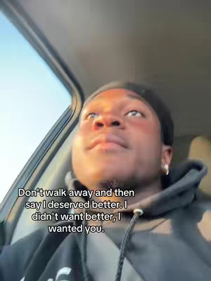 A selfie-style video shot from inside a car. A young Black man is visible from the chest up, looking up and slightly to the side with a thoughtful expression. He is wearing a black hoodie and a black durag. The scene is lit with natural, warm light filtering through the car window, suggesting either sunrise or sunset, casting a soft glow on his face. The car interior is visible, creating a sense of intimacy and immediacy.