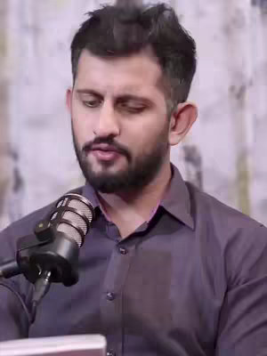A man with a beard and dark wavy hair is positioned slightly left of the frame, looking downwards. He is wearing a dark indigo button-down shirt. A silver microphone is positioned in front of his mouth.