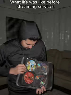 A young man in a black hoodie holds a clear plastic case containing several DVDs. He appears to be looking down at the DVDs. The background is dimly lit, and a glimpse of a window and beige couch is visible.