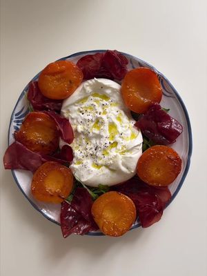 An overhead shot displaying a plate of food. The plate contains a central mound of white cheese topped with olive oil and black pepper. Around the cheese are arranged halves of roasted apricots, slices of what appears to be cured meat, and some green leaves, possibly arugula or a similar leafy green. The plate is white with a blue patterned border, and the background is a plain white surface.