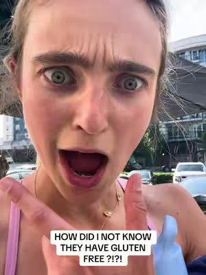 A selfie-style shot with a woman looking directly at the camera with a wide, open-mouthed expression of surprise. Her eyes are dilated, and her eyebrows are raised in a look of shock. The text overlayed on the bottom of the image says, 'HOW DID I NOT KNOW THEY HAVE GLUTEN FREE ?!?!'. The background is blurred, suggesting an outdoor setting.