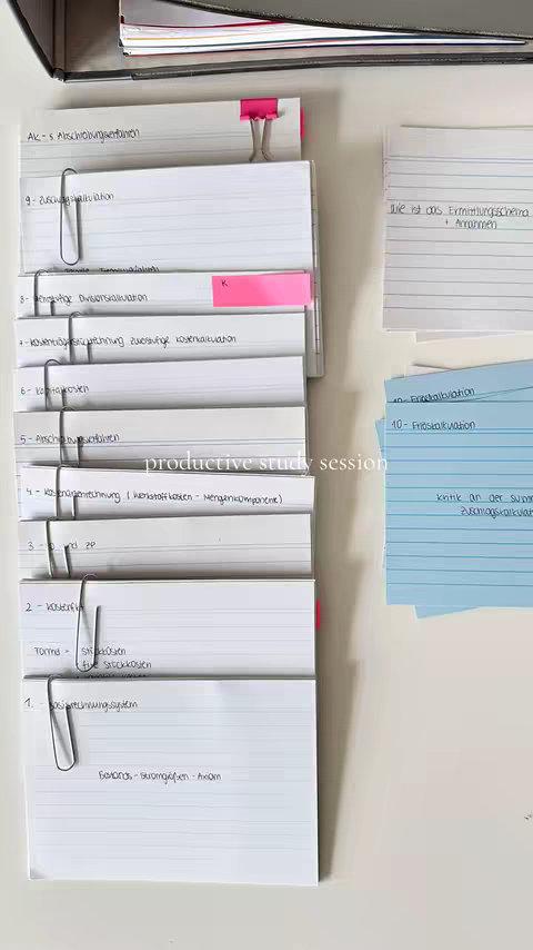 The image shows a vertical stack of flashcards held together with clips and binder. Some have text in German. The hook 'productive study session', is overlaid on the flashcards. Various sheets of lined paper and notepads are also visible. The color palette is muted, with white, shades of blue, and pink. The composition suggests a detailed and organized study setup.