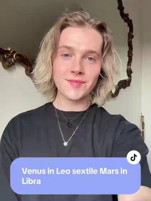 A fair-skinned person with long blond hair stands in front of a blurry brown-colored mirror and wall. The person has a necklace with a crescent and a pendant with a milky-white stone. The person wears a black shirt. A purple graphic square on the bottom has white text.