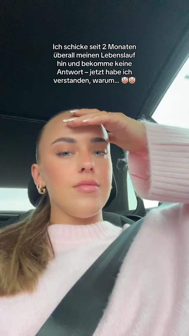 A young woman with fair skin and light brown hair pulled back in a ponytail is in a car. She is wearing a light pink sweater and has a somewhat surprised/exasperated expression, with her hand up near her forehead. The background visible through the car windows is out of focus.