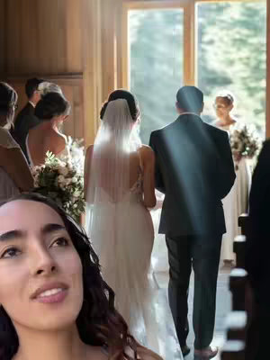 The cover image is a split-screen composition. In the foreground, the close-up of a woman's face is visible. The woman has dark wavy hair and is looking upwards with a subtle smile. In the background, a wedding ceremony is taking place inside a venue or church with large windows. The focus is on the bride, groom, and other members of the wedding party as they proceed down the aisle. The lighting is bright and airy.