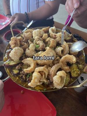 A close-up shot of a dish, featuring a mix of ingredients. The center is filled with what appears to be a stew-like base, topped with crispy, golden-brown chicharon. The dish is served in a large, round, metal dish, with a few hands and utensils visible, suggesting serving. The color palette is a blend of warm browns, yellows, and reds, with contrasting textures of crunchy chicharon and the softer stew base.