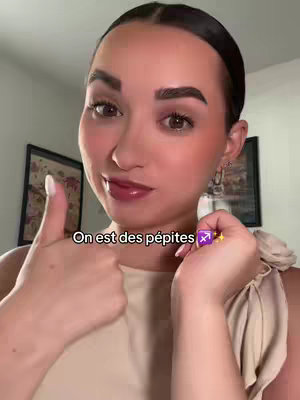 A woman is giving two thumbs up while holding a small object with a Sagittarius symbol on it. Text overlayed reads "On est des pépites". The background shows a room with paintings.