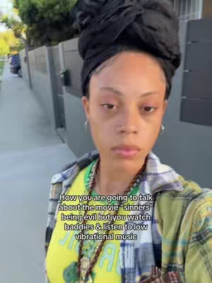 A selfie-style shot of a person standing outside, with a casual outfit and a serious expression. The person has dark hair styled in a bun and is wearing a plaid shirt over a yellow t-shirt. Text overlayed on the image poses a rhetorical question referencing listening to low vibrational music and watching "baddies."