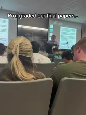 A classroom setting where a professor is standing in front of a screen, likely grading papers, while several students are sitting in the room. The front of a woman with blonde hair in a ponytail is facing to the audience, while others are partially visible.