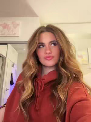 A young woman with long wavy brown hair wearing a red hoodie looks up with a slight smile, a world map decorates the wall behind her. She is in a room that seems to be a bedroom.