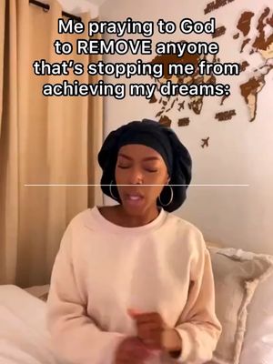 A selfie-style shot featuring a woman in a casual setting. She is wearing a black head covering and a cream-colored sweater. The background includes a curtain and a world map, suggesting a home environment. Text overlay reads 'Me praying to God to REMOVE anyone that's stopping me from achieving my dreams:'