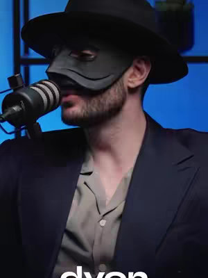 The image features a man wearing a black hat and mask, holding a microphone. The background is blue, and the composition is a medium shot. The man is in the middle of talking.