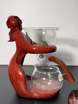 The image showcases a finely crafted tea set supported by a wooden and red structure. A clear glass container holds a small, round stone at the center. The tea set sits on a dark surface. A person's face is partially visible at the lower right corner.