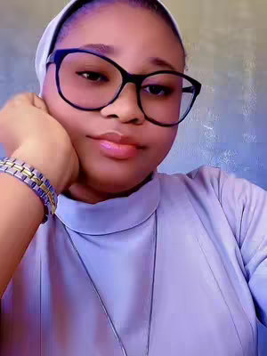 A young woman with light skin and dark hair poses in a close-up shot. She wears large, square-framed black glasses, a white head covering, and a pale blue turtleneck. Her left hand is gently propped against her cheek, and a gold and silver bracelet adorns her left wrist. The background is softly blurred, with a muted gradient of colors.