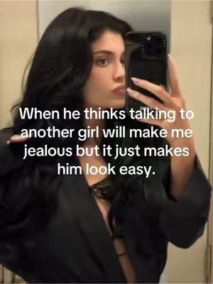 A young woman with long black hair takes a selfie in front of a mirror. She is wearing a dark-colored top and holding a phone in her right hand to take the picture. Text overlayed on the image reads, "When he thinks talking to another girl will make me jealous but it just makes him look easy."