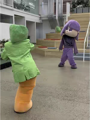 Two individuals dressed as characters from the video game Plants vs. Zombies are positioned in a brightly lit indoor space. One is wearing a green peashooter costume, faces away from the camera on the left, and appears to be in motion. The other is dressed in a purple Lily Pad costume and is looking towards the peashooter with a green throwable object in mid-air between them. The background presents a modern, indoor setting with stairs, and a high ceiling.