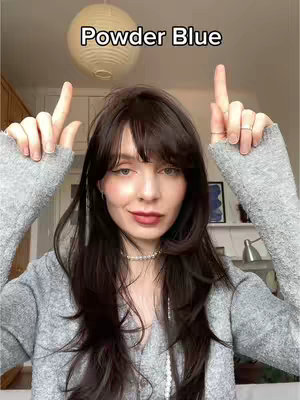 A woman with dark hair and bangs is shown in a close-up shot, pointing upwards with both hands. Above her head, the text "Powder Blue" is displayed. She is centered with a background that has a round, cream-colored light fixture.