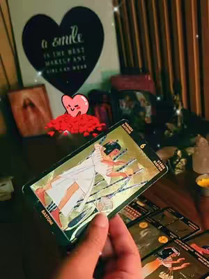 The cover image features a hand holding a tarot card in front of a softly lit, cozy background. The tarot card is the central focus, featuring a woman in white on a gold and black design. In the background, there is a wall decor with a message and a heart, and a soft-focus view of items placed on a wooden table.