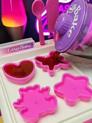 The image shows an Easy-Bake Mix 'N Make Center, with a tray holding various molds and containers. A purple container is pouring a red liquid into the molds, which are in the shape of a heart, a dinosaur, and a star. The background includes a purple lava lamp and other items.