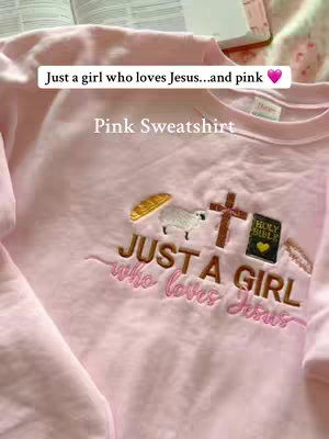 The cover image showcases a pink sweatshirt embroidered with Christian-themed elements. The sweatshirt is laid out on a surface with an open book visible in the background. Text overlays read 'Just a girl who loves Jesus...and pink' and 'Pink Sweatshirt'. The design on the sweatshirt includes a cross, a Bible, a loaf of bread, and a sheep, all aligned in a row.