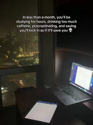 A night scene from a high-rise apartment. A laptop displaying text is open and illuminated on a desk. A tablet and a window with city lights and reflections are also visible. The overlay text is white, and the image is in a dark, moody setting.
