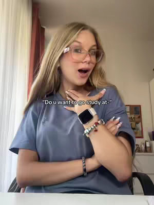A young woman with long blonde hair, wearing glasses and a blue t-shirt, opens her eyes widely with an excited expression. Her arms are crossed, with her hands near her face, showcasing wrist accessories.