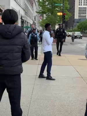 A man wearing a white shirt and black pants speaking into a microphone on a sidewalk. Behind him are multiple individuals, including police officers, and a building with shopfronts. The scene takes place on a city street with pedestrians and vehicles in the background.