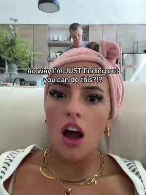 A young woman with a pink turban on her head is the central focus, her face expressing surprise with her mouth open. She is wearing a white top and a gold necklace. Behind her, a man is visible in a domestic setting. Text overlays the image in a white font.