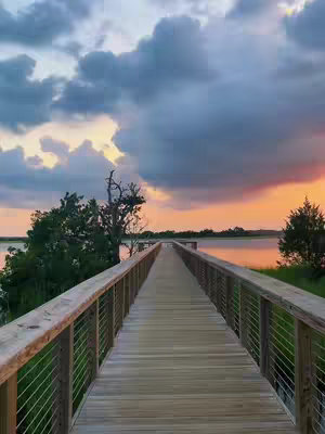 A long wooden boardwalk extends towards the horizon over calm water during dusk. The sky is filled with dramatic clouds, blending shades of blue, grey, and orange from the setting sun. Green vegetation and trees line the sides of the boardwalk.