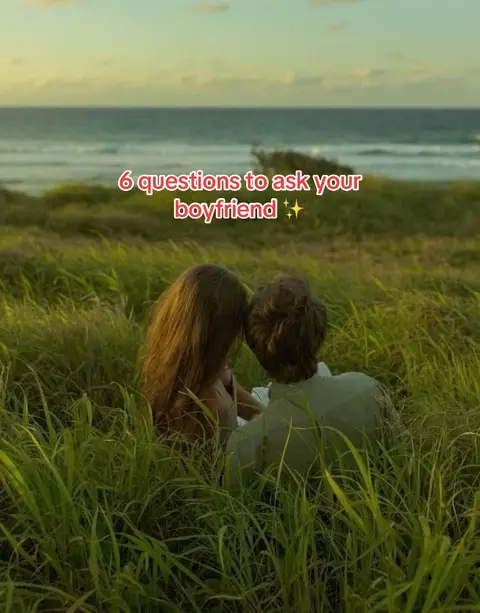 A couple is seated in tall, green grass with each other.  A body of water is visible in the background, and the horizon is clear with a gradient sky and clouds. The image is framed with a vertical aspect ratio.