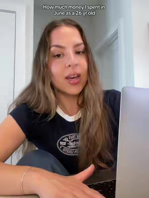 A woman with long brown hair is seated indoors, looking at her laptop. Text overlayed on the image reads, "How much money I spent in June as a 26 yr old". The background appears to be inside a residence or office, likely with white walls and a doorframe visible.